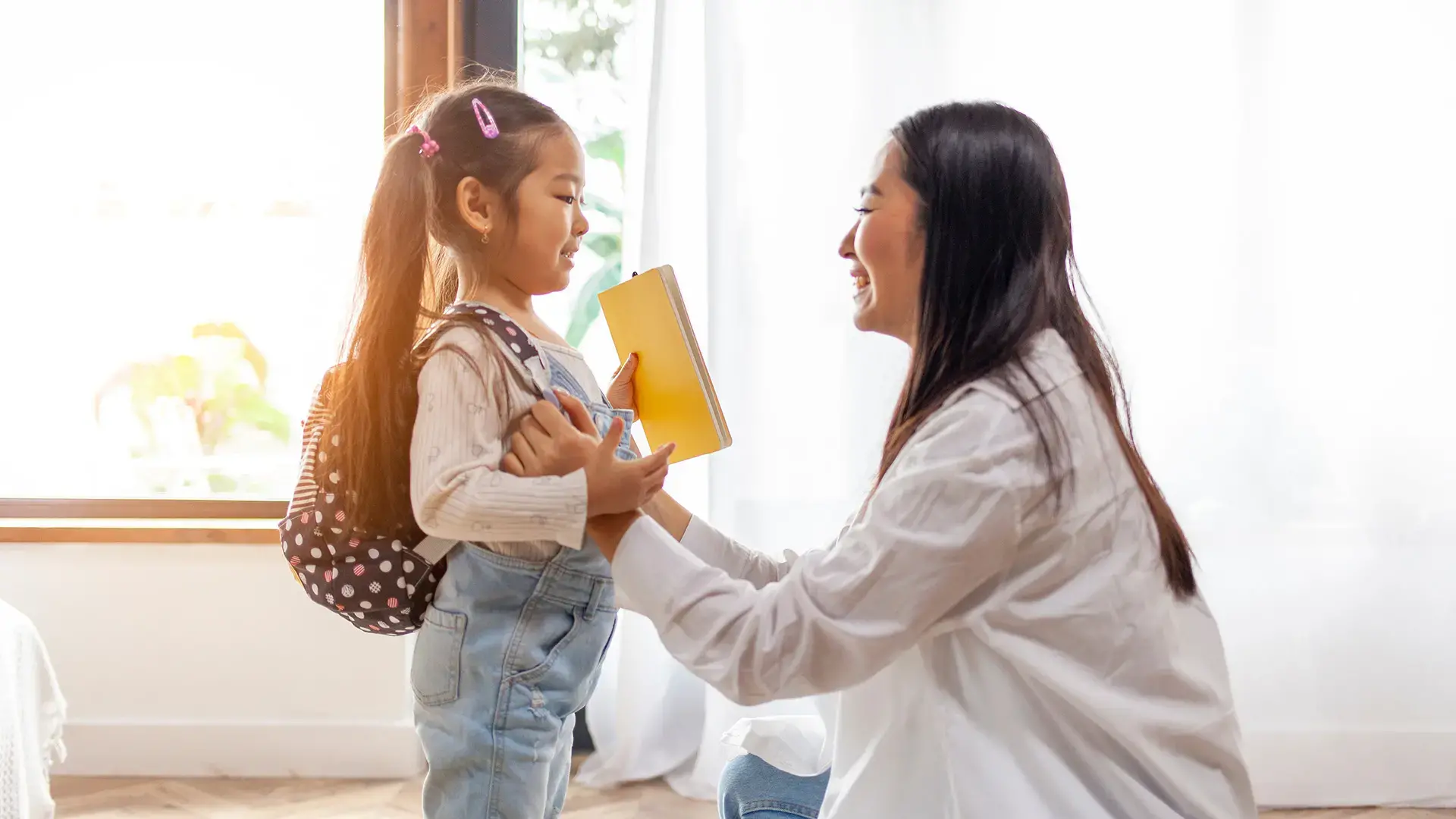 Woman helps her daughter get ready for school and puts her backpack on and supports the girl