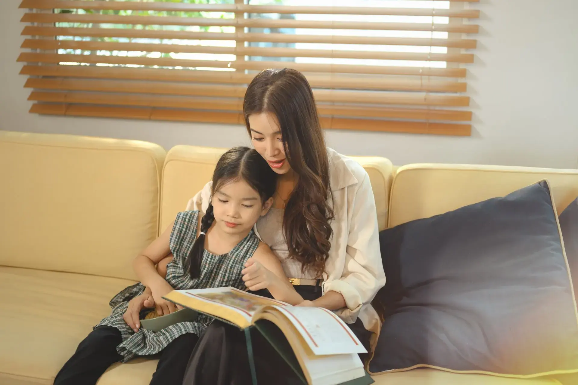 Asyanong nanay at batang anak na babae masayang nagbabasa habang nakaupo sa sofa.