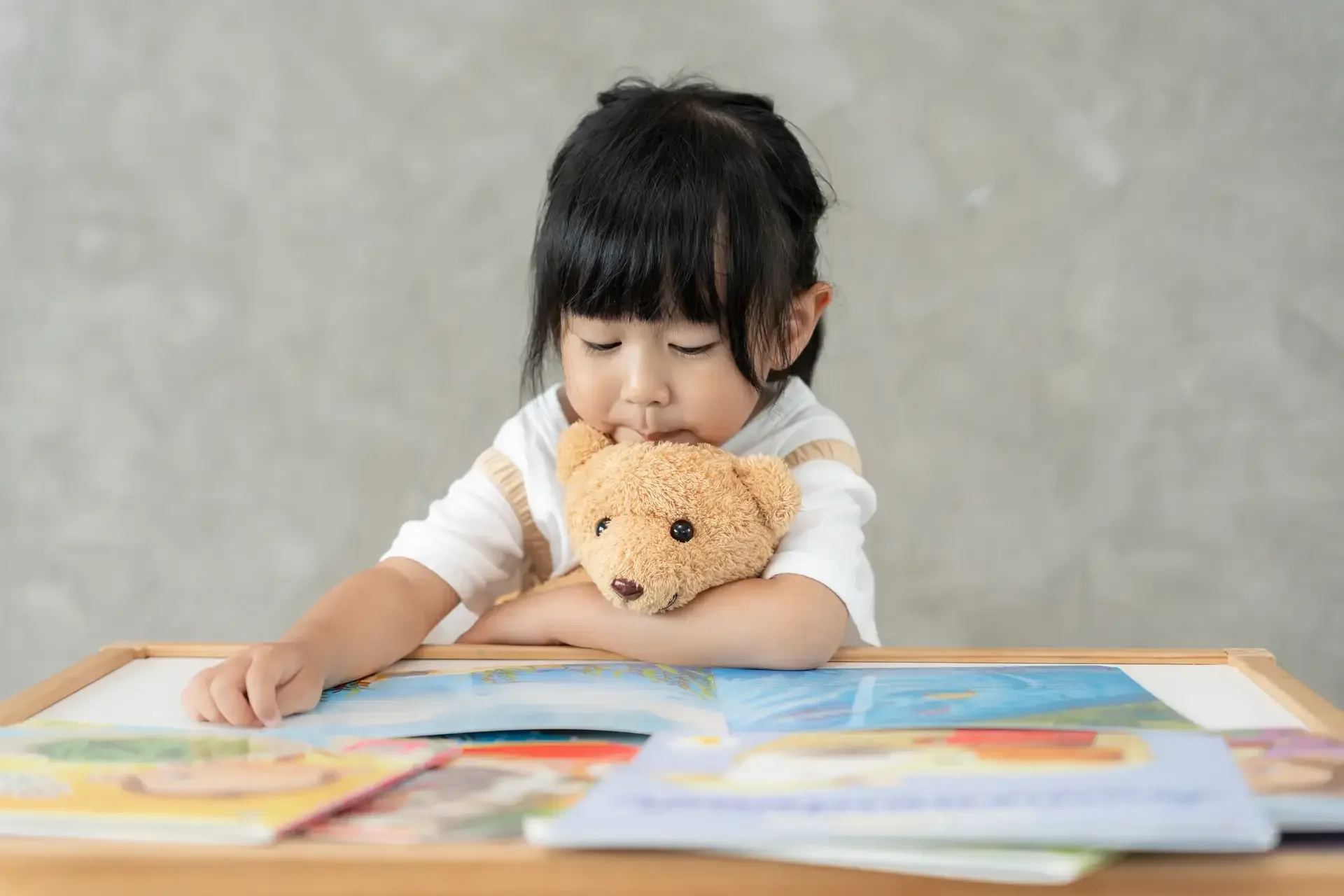 Asyanong batang babae na nagbabasa ng mga libro habang may yakap na teddy bear.